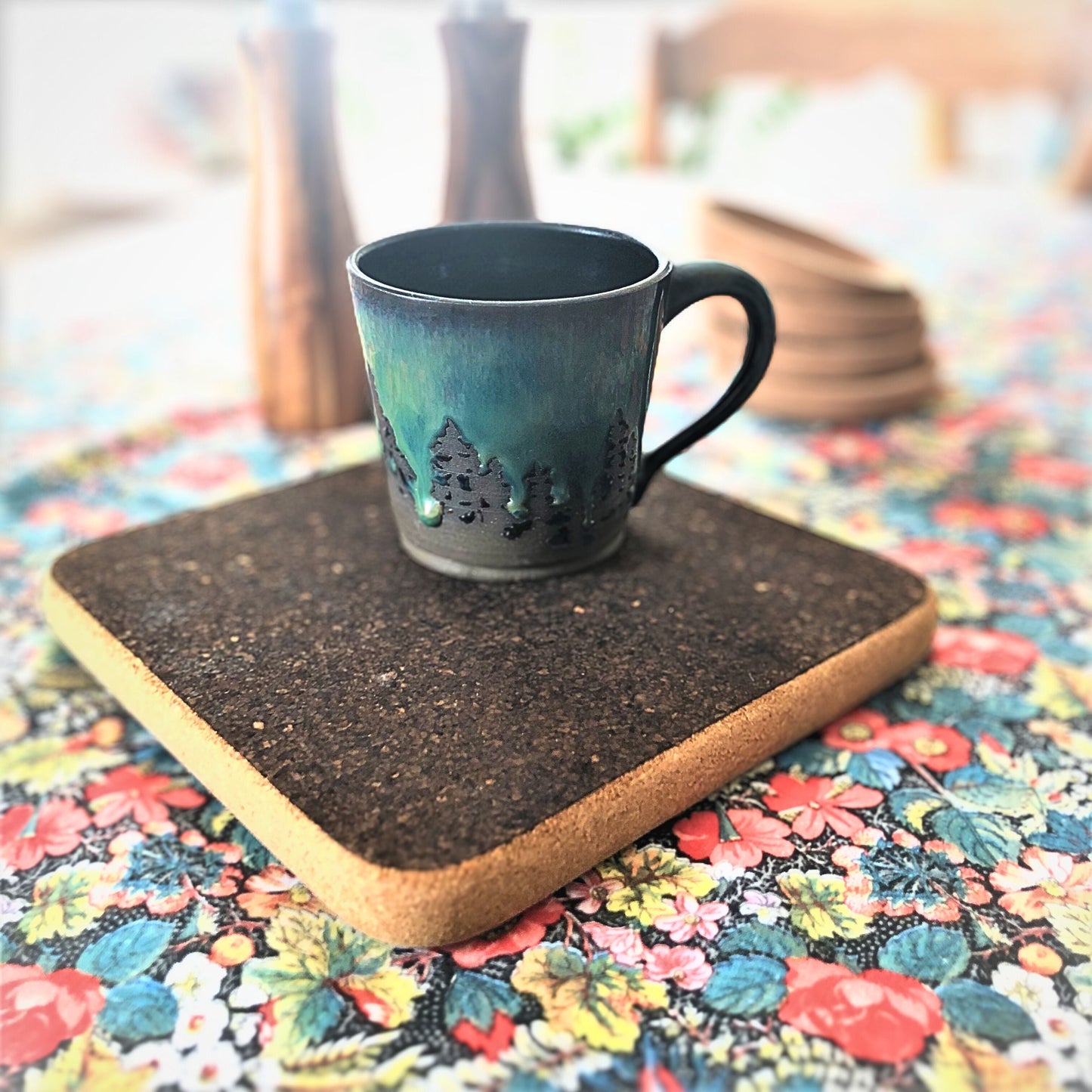 A dark cork sandwich on a colorful floral table cloth with a handmade ceramic black mug with tonal green glaze mimicking the night sky over a pine forest. 