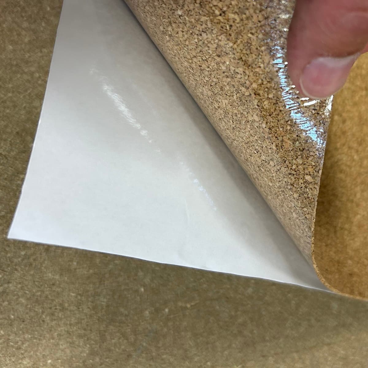 Close-up of a cork sheet with a piece of white paper peeled back showing the PSA backing, held by a hand.